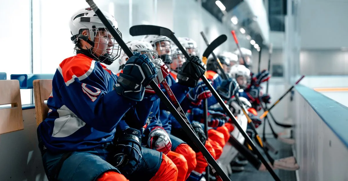 Young ice hockey players in uniforms sitting on the bench during a game, holding hockey sticks.