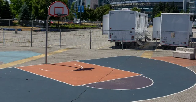Outdoor basketball court in urban setting, sunny day, city skyline and stadium visible.