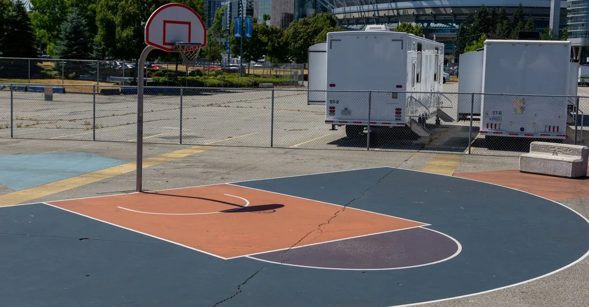 Outdoor basketball court in urban setting, sunny day, city skyline and stadium visible.