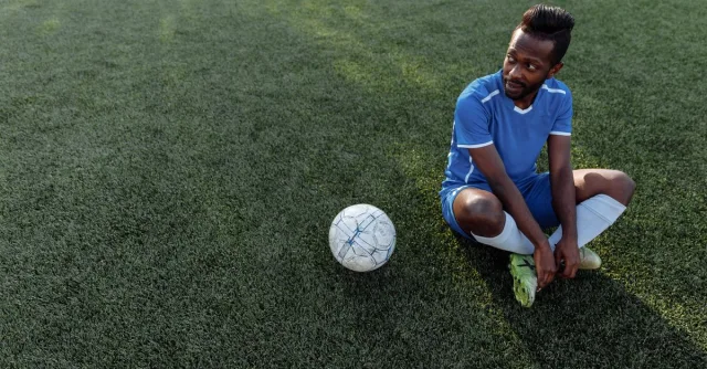 A soccer player in a blue jersey sits on the grass field during the day.