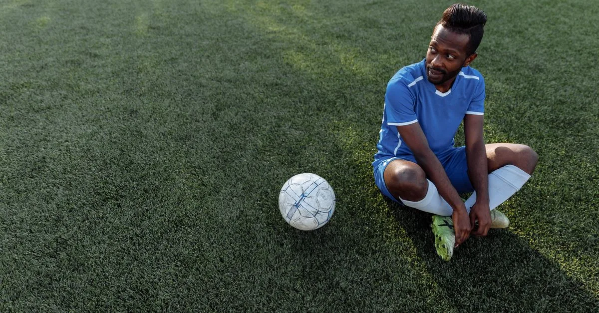A soccer player in a blue jersey sits on the grass field during the day.