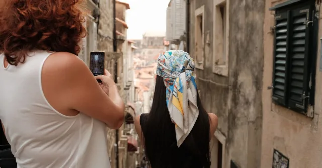 Two women explore a narrow alley in Dubrovnik's historic old town, capturing the moment on a smartphone.