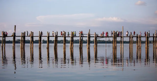 People walking on the iconic U Bein Bridge, with a serene water reflection in Amarapura, Myanmar.