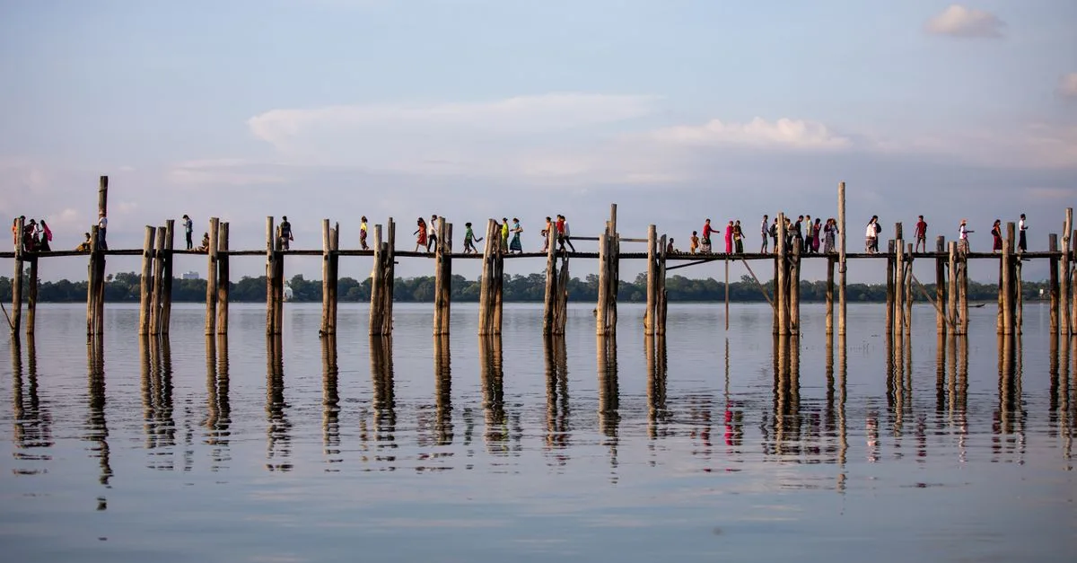 People walking on the iconic U Bein Bridge, with a serene water reflection in Amarapura, Myanmar.