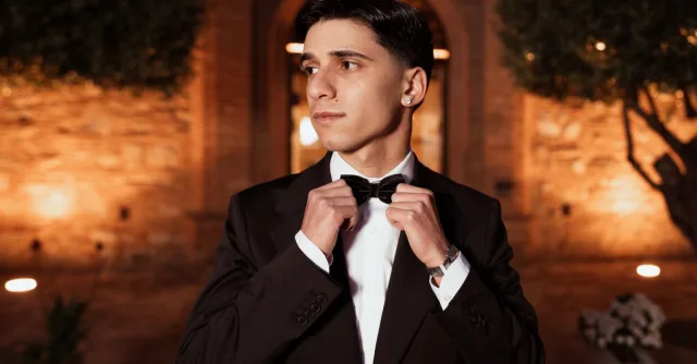 A young man dressed in a tuxedo adjusting his bow tie outdoors at night in Italy, creating an elegant and sophisticated ambiance.