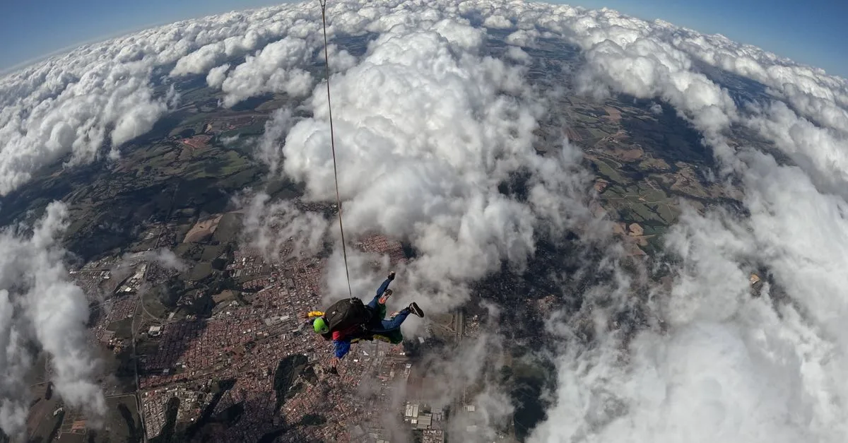 A thrilling aerial shot of a skydiver under parachute above a city and clouds.