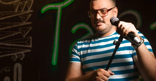 Man holding a microphone doing stand-up comedy in casual striped shirt on stage.