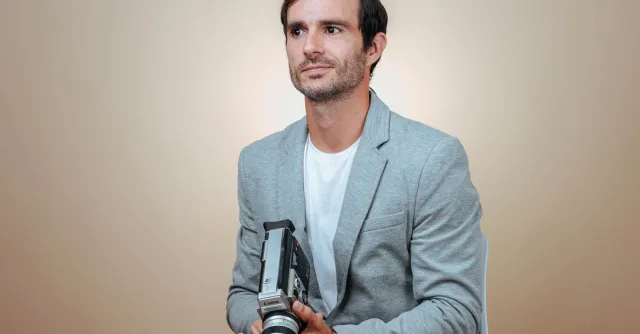 Man sitting with a vintage camera against a beige background in studio lighting.