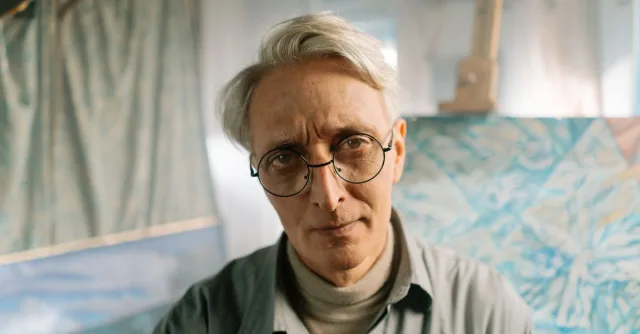 Portrait of a senior artist in glasses at his studio, surrounded by abstract art.