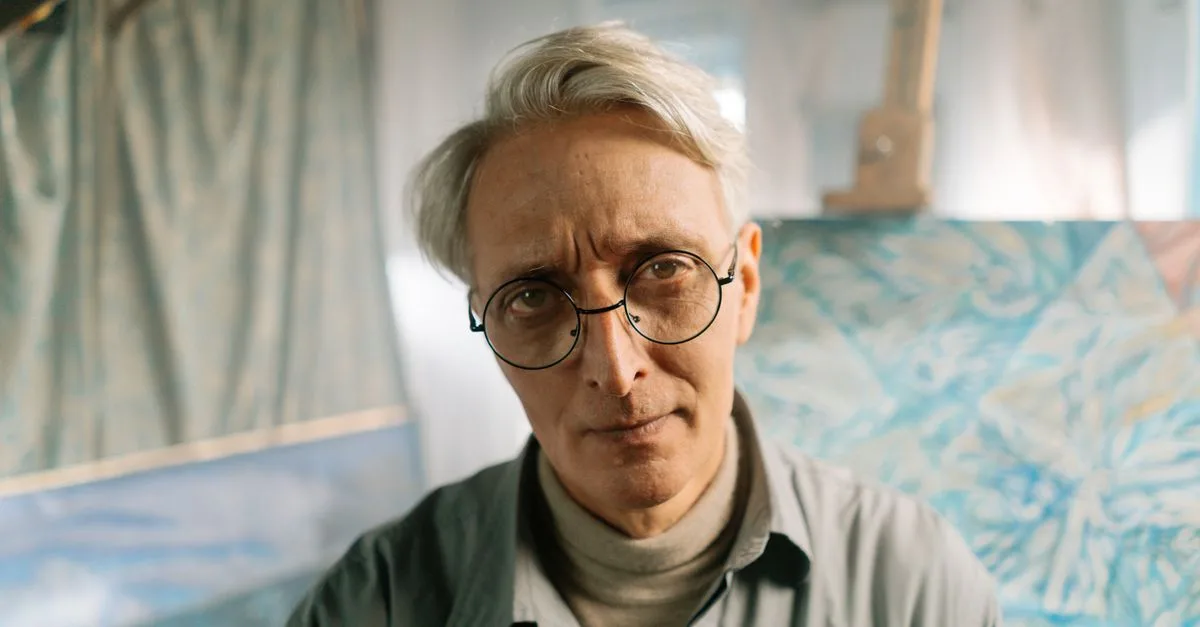 Portrait of a senior artist in glasses at his studio, surrounded by abstract art.