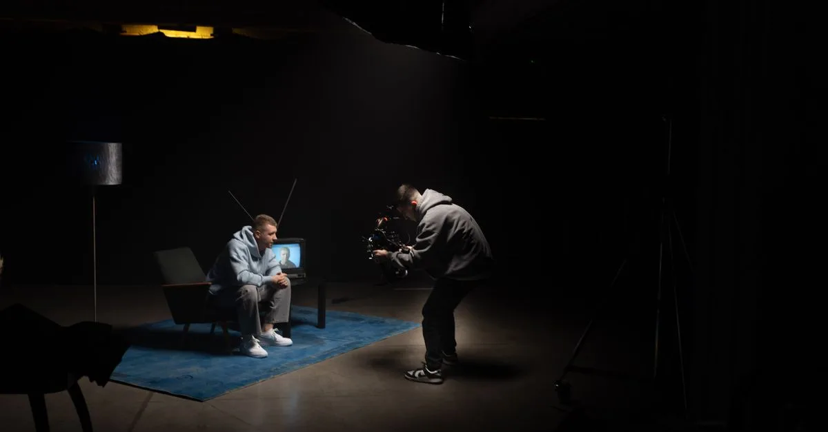 Film production shoot indoors with cinematographer filming an actor sitting, studio lighting.