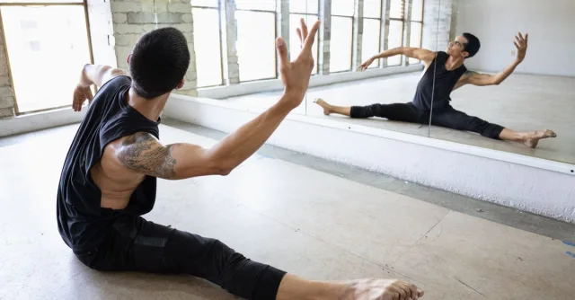 A male dancer performing a stretch in a dance studio with mirrored reflection.