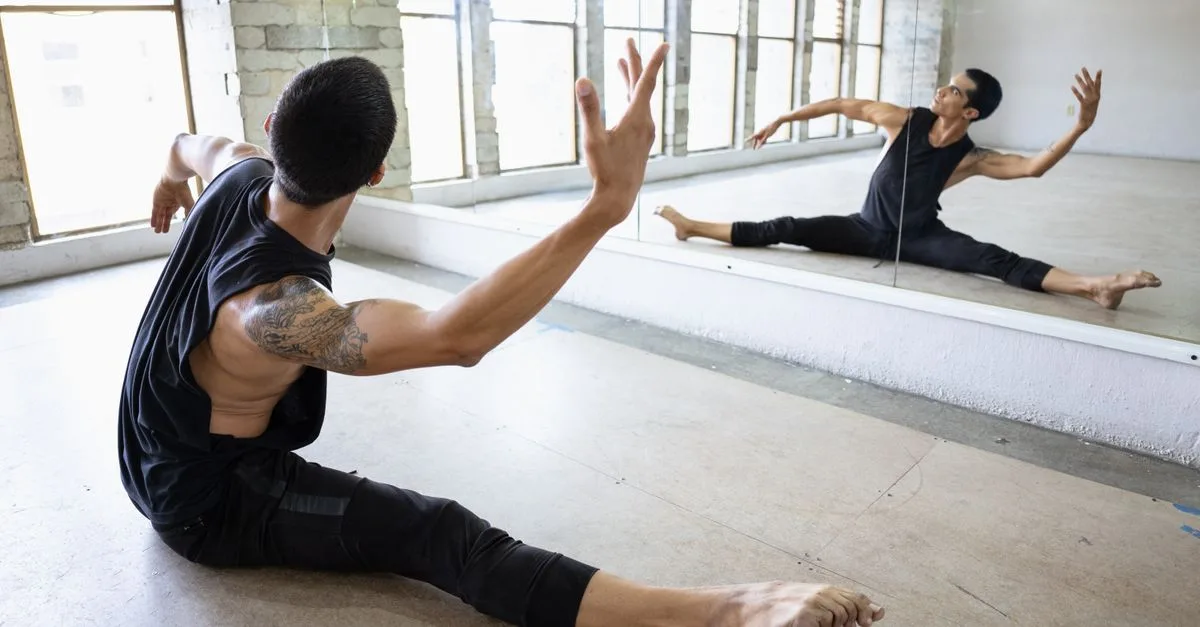 A male dancer performing a stretch in a dance studio with mirrored reflection.