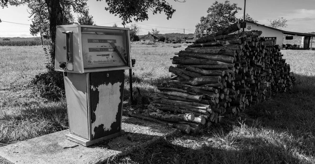 Black and white image of an old gas pump beside a woodpile in the countryside.