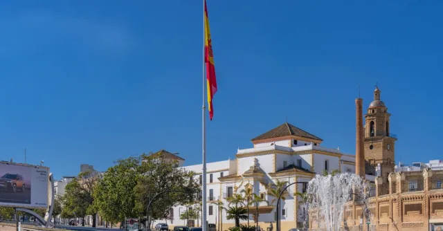 Scenic view of a plaza in Cádiz with a prominent Spanish flag, a fountain, and historic architecture.