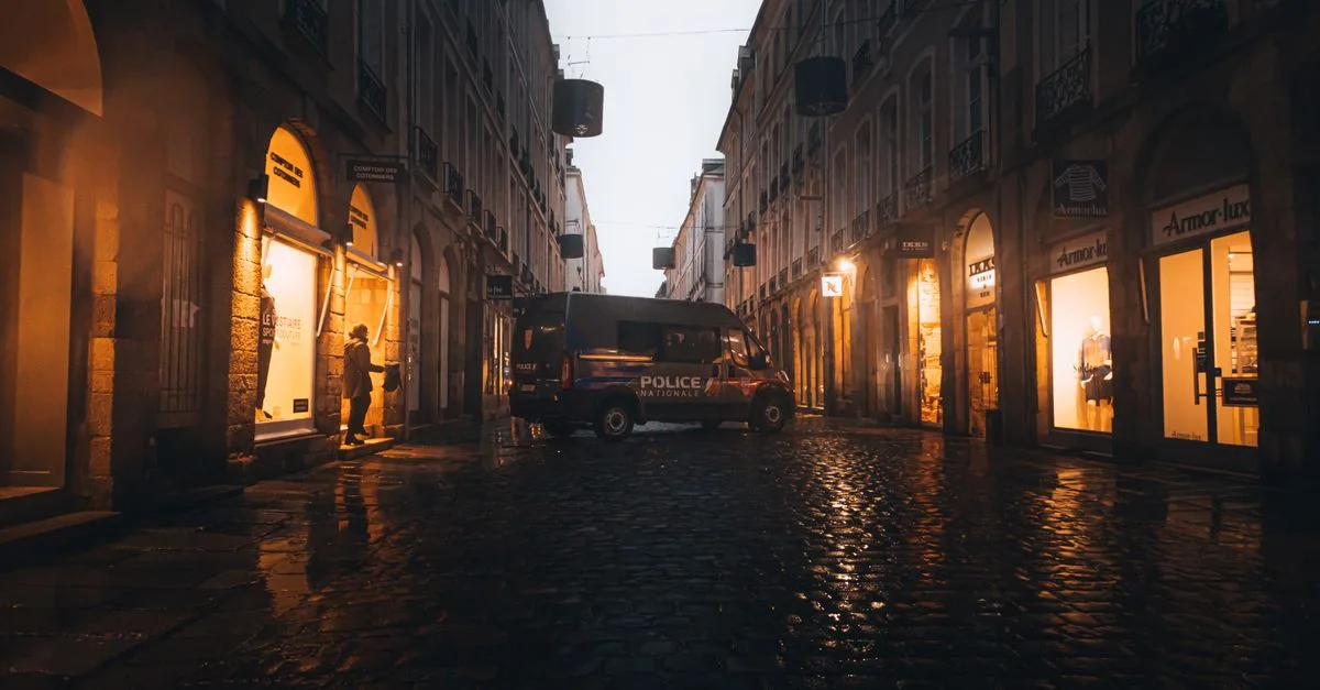A dimly lit city street with a police van and a person illuminated by storefronts at twilight.