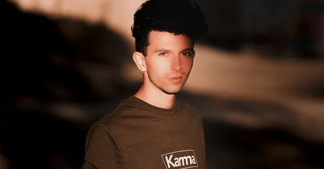 Portrait of a young man with black hair wearing a brown blouse with 'Karma' text, standing outdoors.