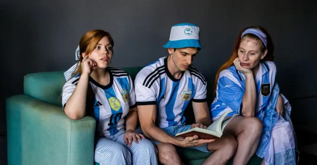 Three adults in Argentina soccer jerseys reading a book indoors on a couch.