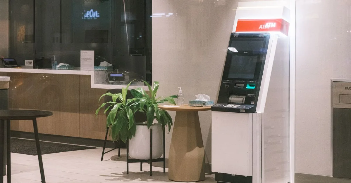 An ATM machine stands in a modern bank lobby, next to a plant and table.