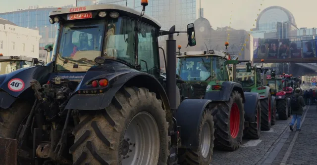 Line of tractors on city street amidst protest with urban buildings in background.