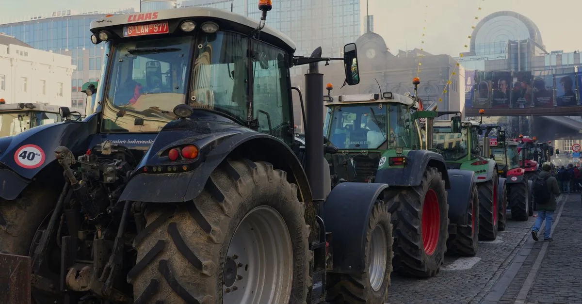 Line of tractors on city street amidst protest with urban buildings in background.