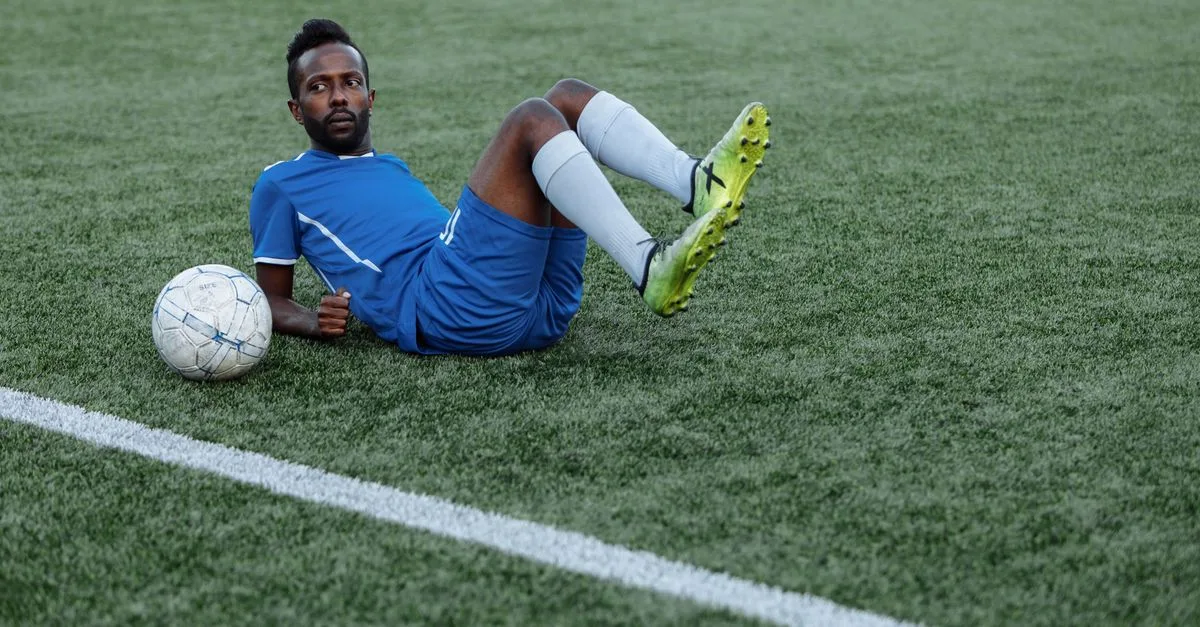 A soccer player in blue jersey lying on the field with a ball beside him.