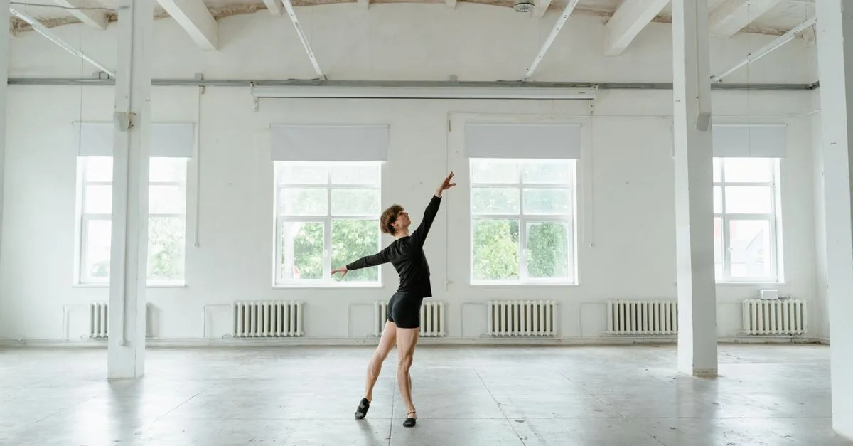 A graceful dancer strikes a pose in a sunlit, spacious studio with large windows.