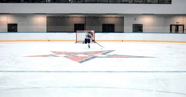 A hockey player in full gear takes a shot on goal in an indoor ice arena.