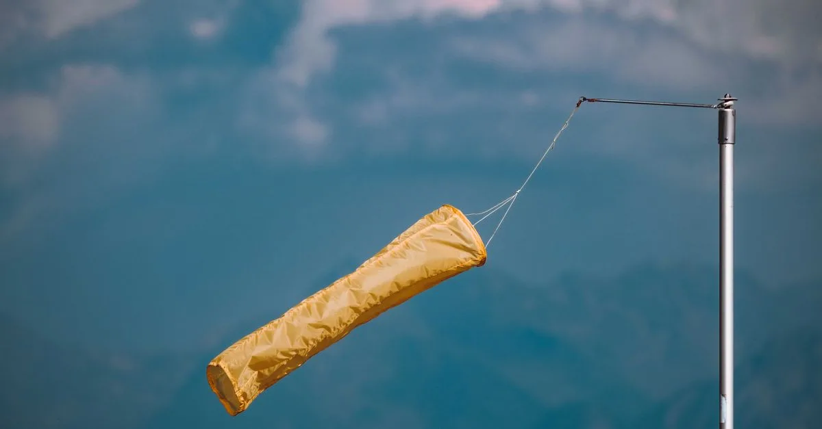 Yellow windsock blowing in the wind, mounted on a pole against a cloudy blue sky.