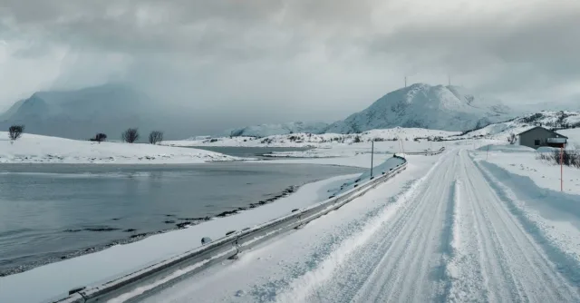 Snow-covered road in Norway with mountains and a lake under cloudy skies.