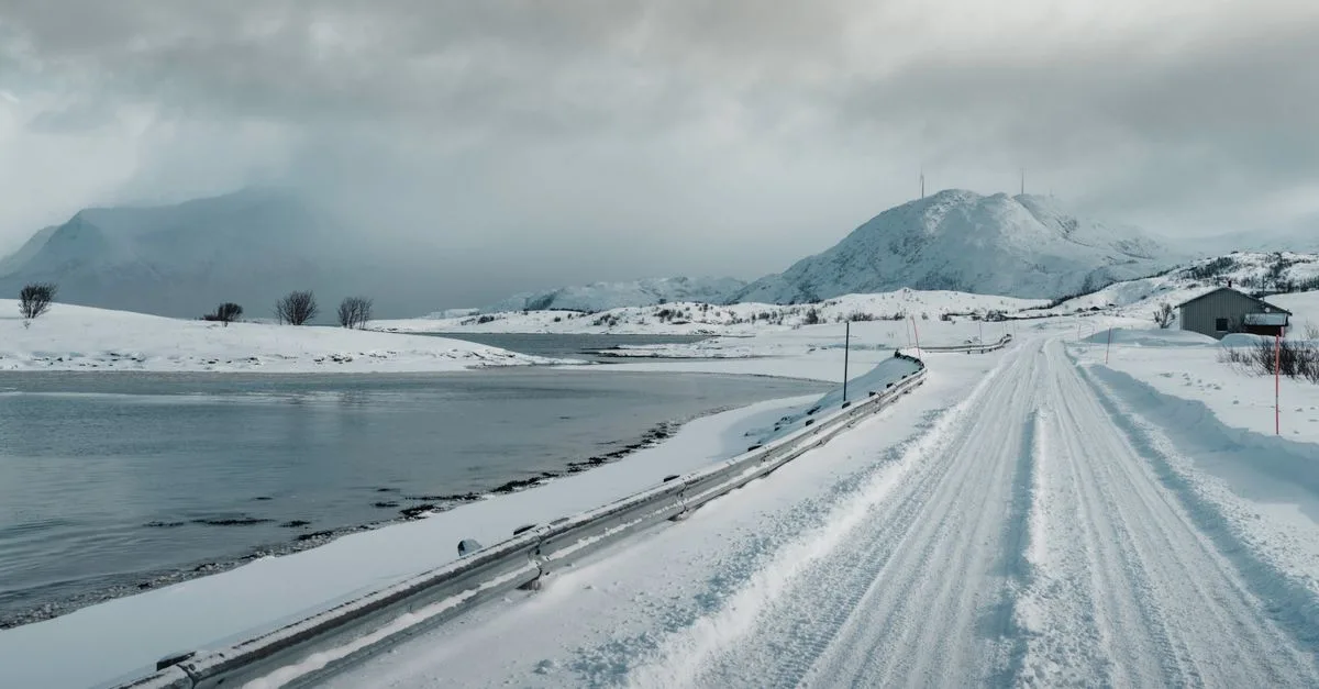 Snow-covered road in Norway with mountains and a lake under cloudy skies.