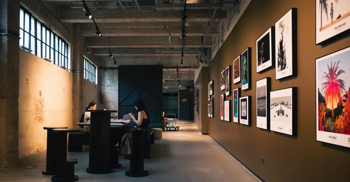Art gallery hallway with framed artworks and visitors seated inside, under warm lighting.