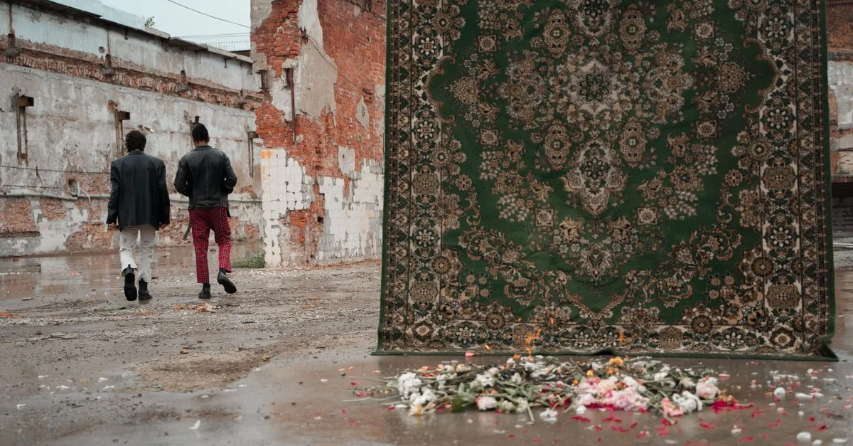Two men walk past a hanging rug and scattered flowers in an abandoned building.