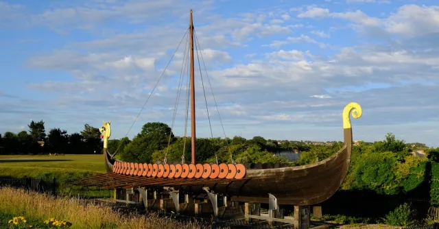 A detailed Viking longship replica set in a green outdoor landscape under clear blue skies.