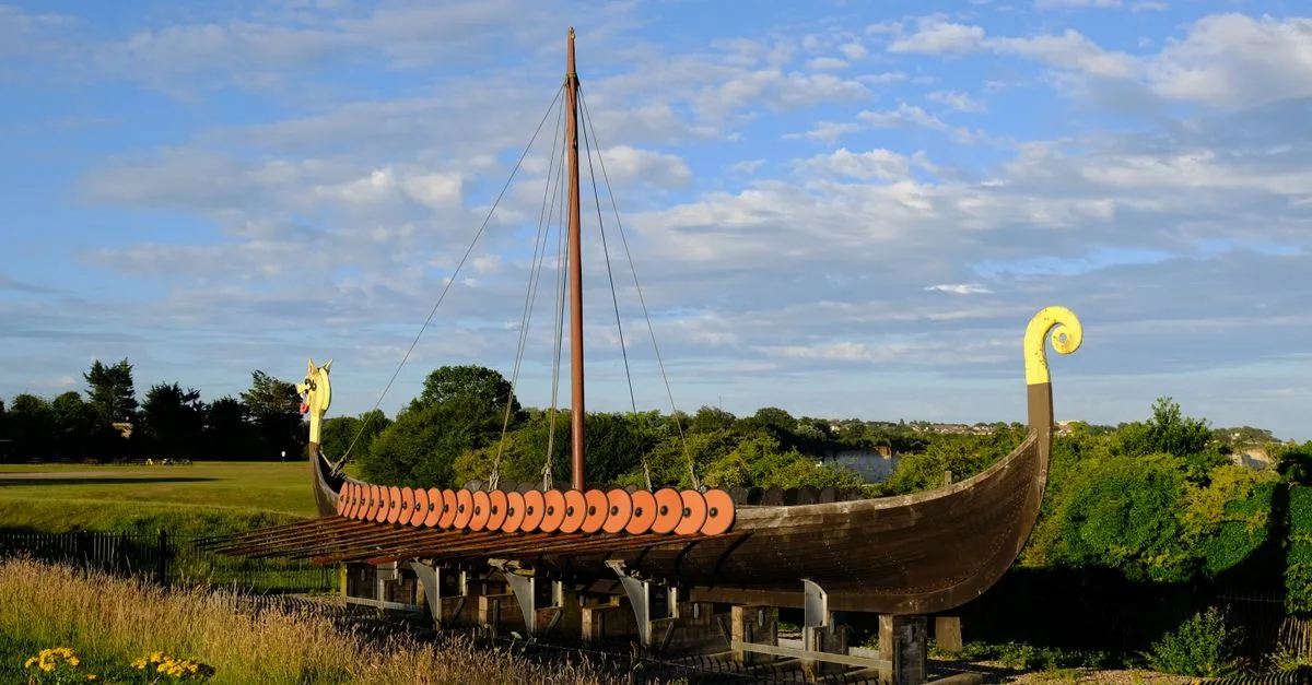 A detailed Viking longship replica set in a green outdoor landscape under clear blue skies.