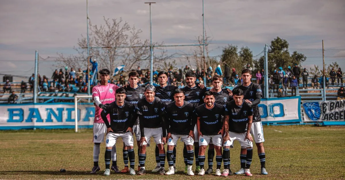 A soccer team poses together on an outdoor field before a match, with spectators in the background.