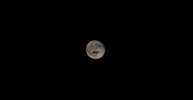 An airplane silhouette against a bright full moon in a clear night sky, showcasing travel and exploration.