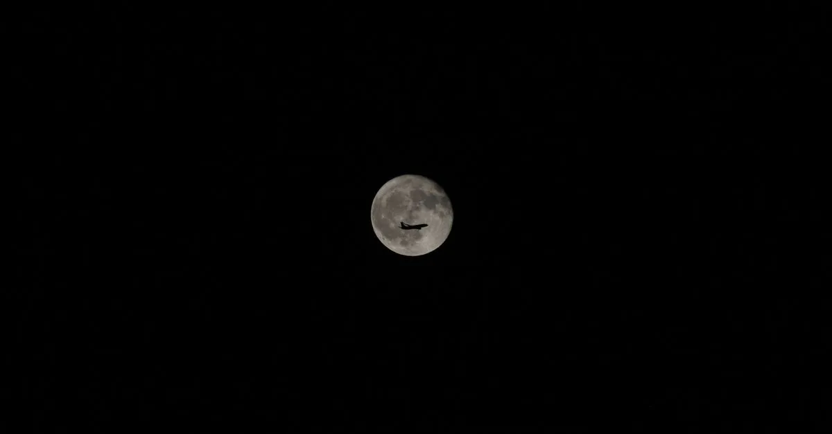 An airplane silhouette against a bright full moon in a clear night sky, showcasing travel and exploration.