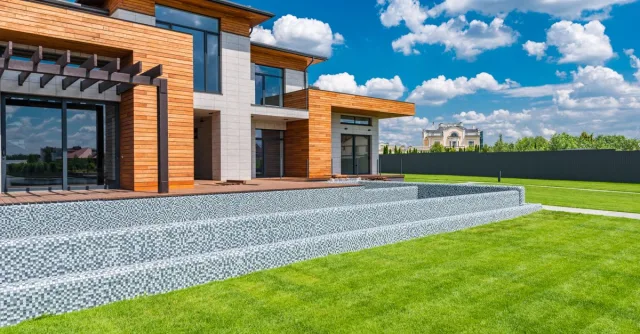 Exterior of contemporary residential house with panoramic windows glass doors and green lawn in yard on sunny day against blue sky with white clouds