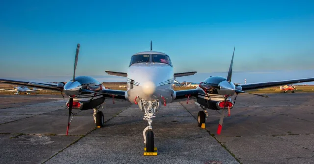 A front view of a twin-engine aircraft on tarmac under clear blue skies.