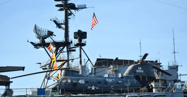 A US Navy helicopter is stationed on the deck of an aircraft carrier with radar equipment in the background.