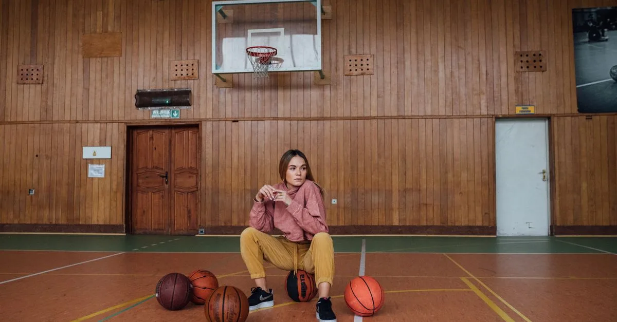 A young woman poses confidently in an indoor basketball court, surrounded by basketballs.