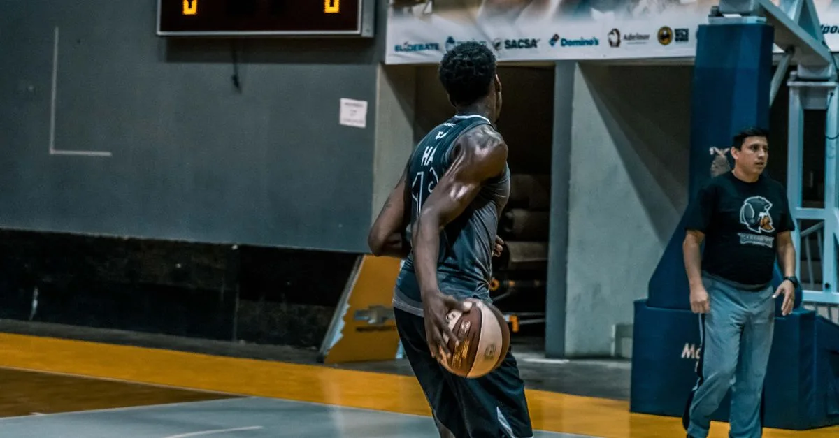 Basketball player performing on an indoor court with scoreboard in background, highlighting athleticism and teamwork.