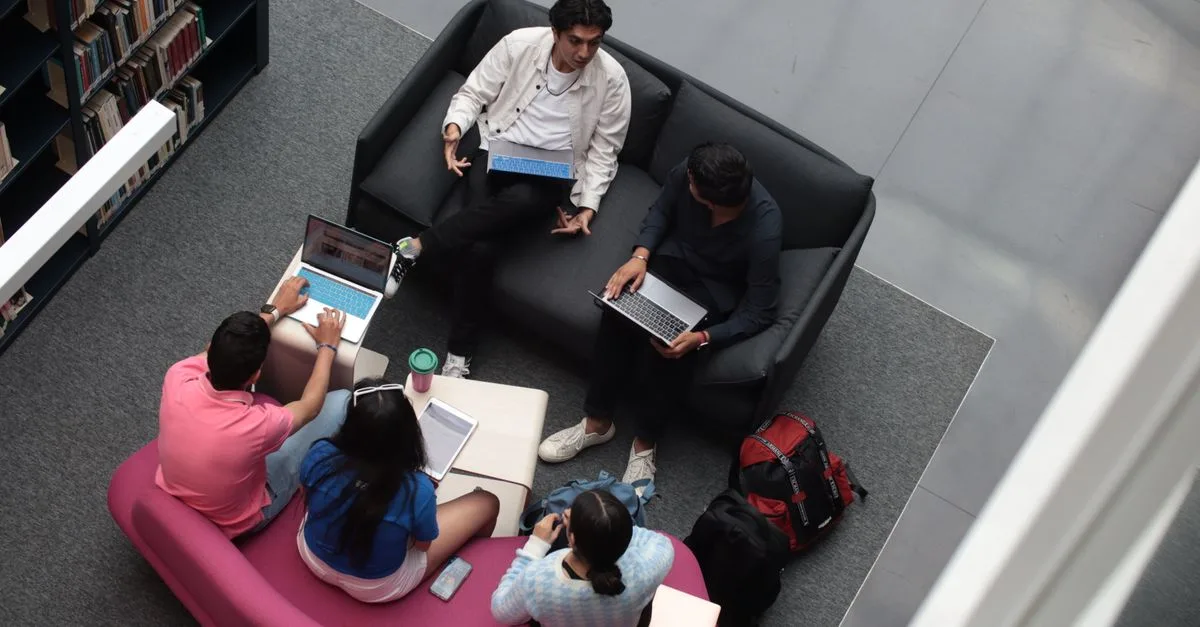 A group of diverse students using laptops and books in a library setting, studying together.