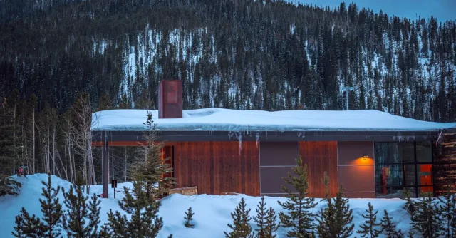 A modern cabin in a snowy forest at twilight, surrounded by mountains.