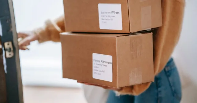Crop anonymous female wearing casual outfit carrying carton parcels with address labels opening door of post office