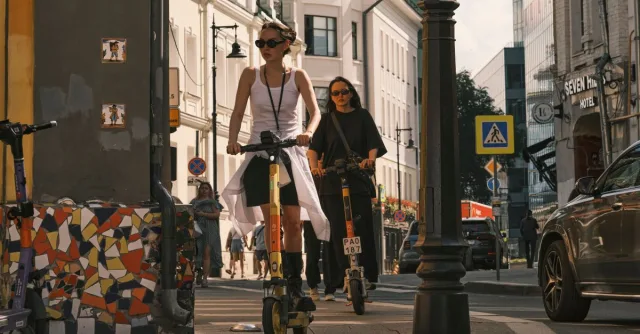 Two women riding electric scooters on a city street, showcasing urban commute.
