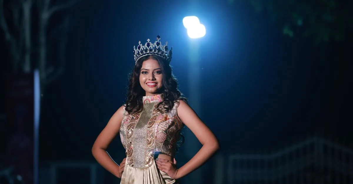 Beautiful woman wearing a crown and gown, posing confidently at night under dramatic lighting.