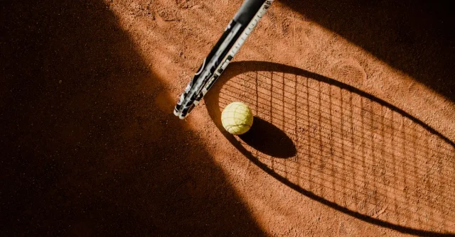 Tennis ball with racket shadow on a sunlit clay court.