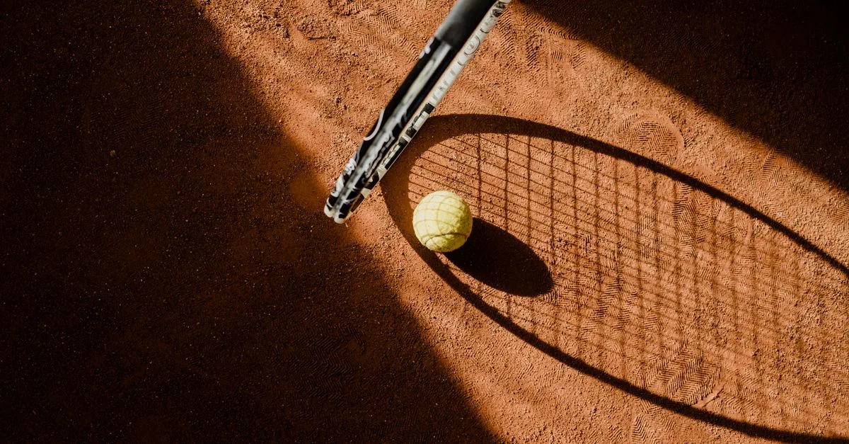 Tennis ball with racket shadow on a sunlit clay court.